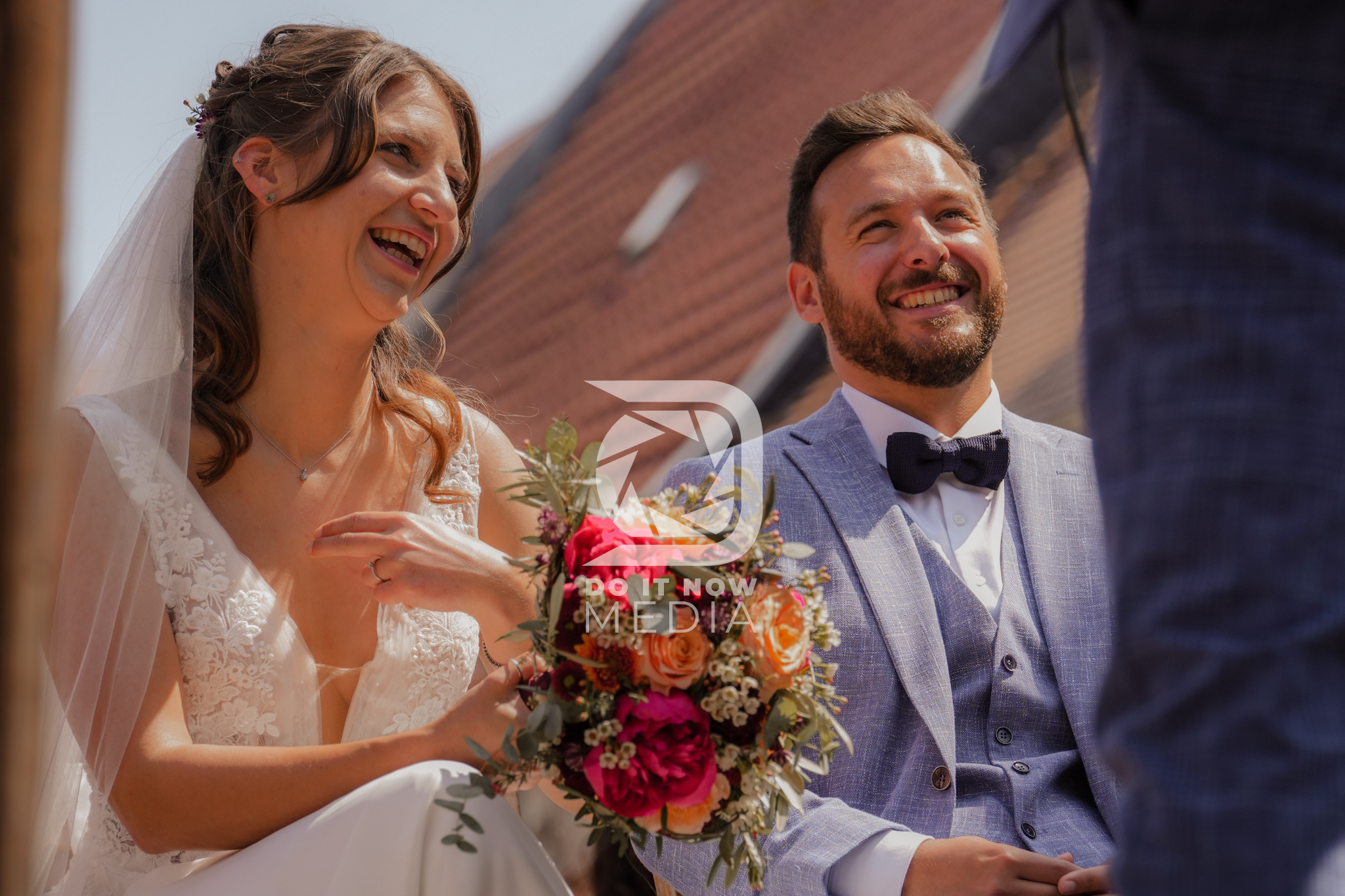 Lisa und Flo lachen während ihrer Trauung unter freiem Himmel, Lisa hält ihren bunten Brautstrauß.